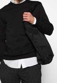Sudadera negra con puños de canalé, llevada sobre una camisa blanca con cuello, combinada con pantalones de rayas en blanco y negro y un bolso negro.