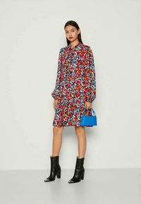 Vestido floral con cuello alto, mangas largas abullonadas y dobladillo acampanado. Patrón multicolor sobre un fondo oscuro, combinado con un bolso azul y botas negras.