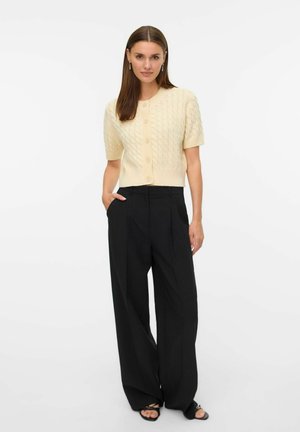 Vrouw in een crèmekleurige cardigan met korte mouwen van kabelgebreide stof, zwarte wijde broek en zwarte sandalen, staand met één hand in de zak.