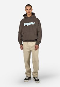 Barna kapucnis felső világoskék "pegador" logóval, kenguruzsebbel, bordázott aljjal, bézs nadrággal és fekete sportcipővel párosítva.