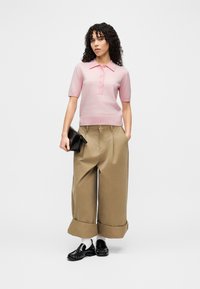 Ροζ πόλο με κοντά μανίκια και κουμπιά, σε συνδυασμό με μπεζ παντελόνι φαρδύ, ψηλόμεσο και μαύρα μοκασίνια, κρατώντας μια μαύρη τσάντα φάκελο.