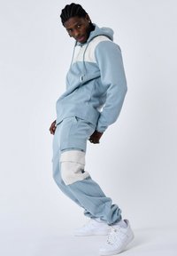 Felpa blu chiaro con pannello bianco sulle spalle, con cordino e tessuto testurizzato; pantaloni coordinati con accenti bianchi e tasche, indossati con sneakers bianche.