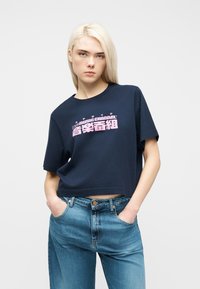 Ξανθιά γυναίκα που φοράει σκούρο μπλε κοντό μπλουζάκι με ροζ κείμενο "EDWIN MUSIC CHANNEL" και μπλε τζιν, με τα χέρια στις τσέπες, ουδέτερη έκφραση.