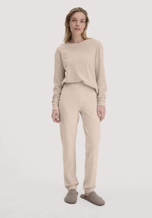 Beigefarbenes Loungewear-Set aus weichem Stoff, bestehend aus einem langärmligen Oberteil und schmal geschnittenen Hosen, dazu passende graue Hausschuhe.
