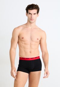 Schwarze Boxershorts mit rotem Elastikbund, der das Logo "HUGO" trägt. Glattes Material, enganliegendes Design und mittelhoher Schnitt.