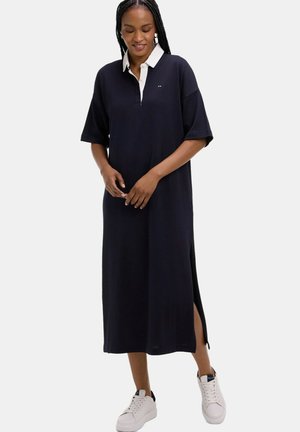 Femme portant une robe midi ample bleu marine avec un col blanc, une fente latérale et des baskets blanches, debout devant un fond uni.