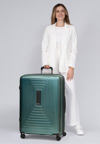 Vrouw in witte outfit staand en een handvat vasthoudend van een grote groene koffer met wieltjes op een grijze achtergrond.