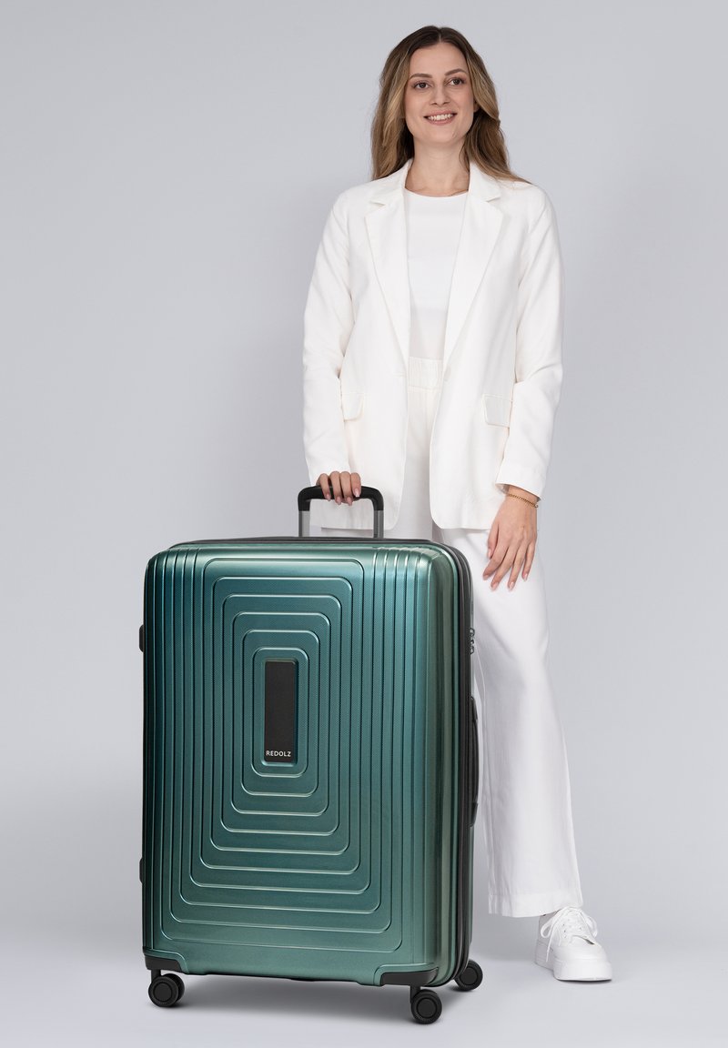 Vrouw in witte outfit staand en een handvat vasthoudend van een grote groene koffer met wieltjes op een grijze achtergrond.