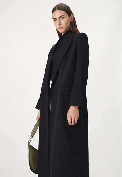 Frau mit glattem braunem Haar, die einen langen schwarzen Mantel und ein schwarzes Oberteil trägt, eine olivgrüne Handtasche hält und vor einem weißen Hintergrund steht.
