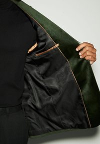 Grüne Wildlederjacke mit schwarzem Netzfutter, die eine Tasche mit Satinbesatz aufweist und geöffnet gehalten wird, um das Innendesign und die Materialien zu präsentieren.