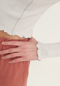 Mano reposando en la cadera, vistiendo una blusa de manga larga ribeteada color blanco roto y pantalones de talle alto en un tono óxido con una cintura fruncida.