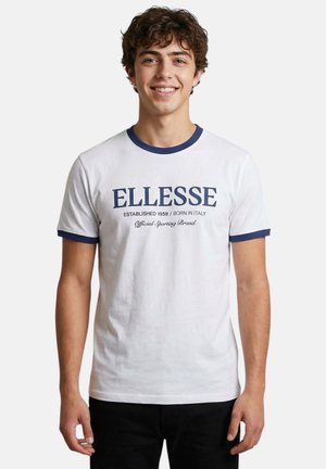 Jeune homme souriant aux cheveux bouclés portant un t-shirt blanc avec bordure bleue et le texte "ELLESSE Established 1959 Born in Italy".