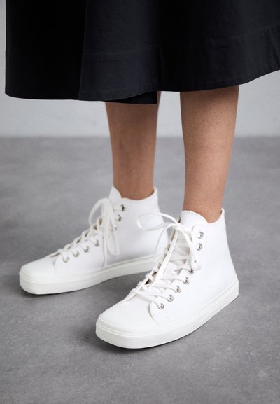 Vita hög-top sneakers i slätt material, med metallögon, platta vita sn�örer och rundad tå. Texturerad sula är synlig.