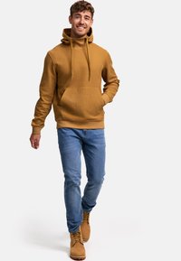 Un hoodie texturé jaune moutarde avec une poche avant, associé à un jean bleu et des bottes à lacets marron. Il comprend une capuche avec un cordon.