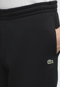 Pantalon de survêtement noir avec taille élastique et poche latérale, avec un petit logo crocodile vert près de la poche.