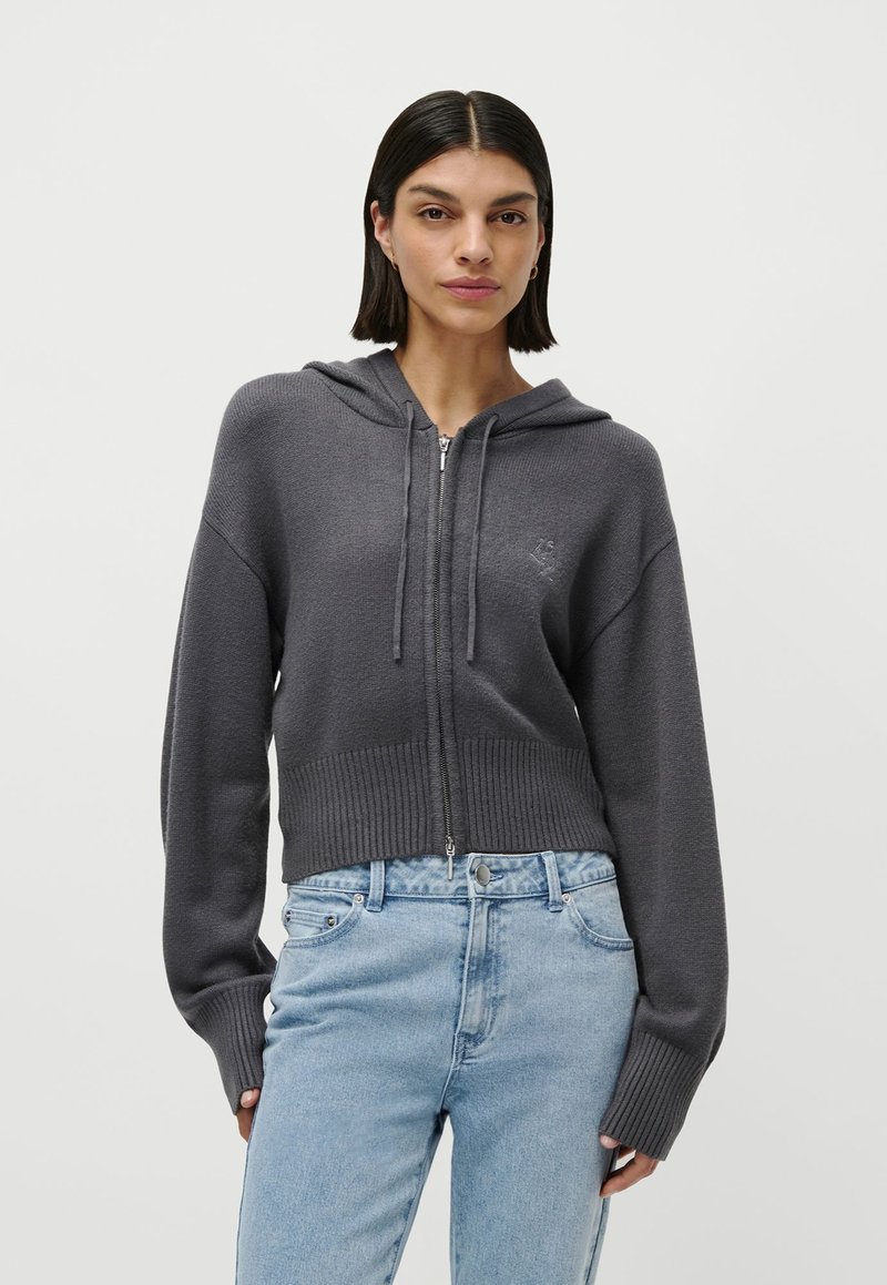 Mujer que lleva un suéter con capucha y cremallera de color gris oscuro y unos vaqueros de tiro alto de color azul claro, de pie contra un fondo liso.