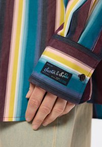 Πολύχρωμο ριγέ πουκάμισο με συνδυασμό τιρκουάζ, μπορντώ, κίτρινο και ροζ. Περιλαμβάνει ετικέτα λογοτύπου και κουμπί στον καρπό.