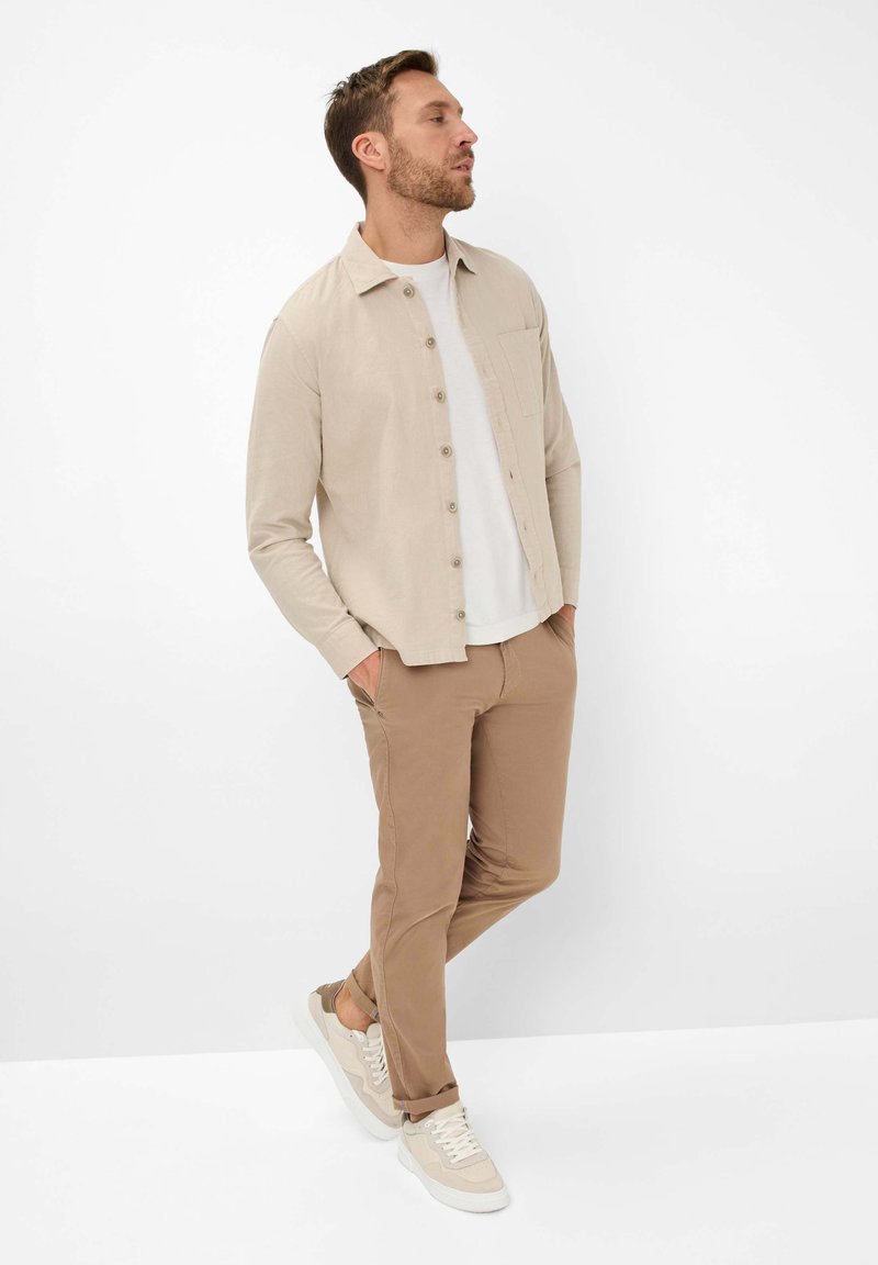 Beige button-up shirt over white t-shirt, paired with beige pants and light sneakers. The shirt features a chest pocket and soft texture.