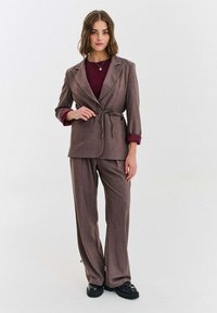 Žena stojaca, oblečená v hnedom obleku s opaskom s uzlom cez bordový top a čierne masívne topánky na jednoduchom bielom pozadí.