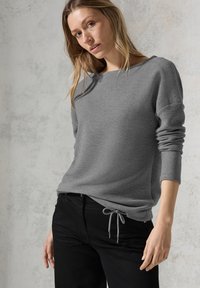 Grijze pullovertrui van een textiele stof, met een brede halslijn en opgerolde mouwen. In combinatie met zwarte jeans met een trekkoordsluiting.