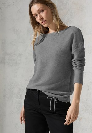 Pull gris à texture, avec un col large et des manches retroussées. Assorti à un jean noir avec une fermeture à cordon.