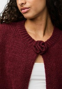 Bordeaux gebreide cardigan met een textuurafwerking en geribbelde kraag. Voorzien van een bloemenvormige sluiting bij de hals en een open voorkant ontwerp.