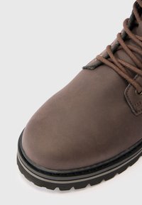 Primer plano de una bota de cuero marrón con suela negra gruesa y cordones marrones a juego sobre un fondo blanco.