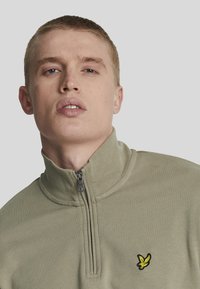 Svetlozelená mikina s vysokým golierom a polovičným zipsom. Obsahuje malé čierne a žlté vyšívané logo na ľavej hrudi.