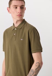 Olivengrøn polo t-shirt med klassisk krave, korte ærmer, tre-knaps lukning og et lille broderet logo på brystet.