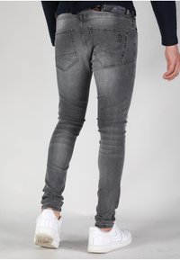 Vaqueros skinny de mezclilla gris con un acabado desgastado, que presentan dos bolsillos traseros y un parche de logo negro en la cintura. Combinados con zapatillas blancas.