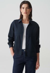 Navy blue button-up shirt with a collar, long sleeves, chest pockets, and a relaxed fit, paired with dark jeans. Smooth fabric texture.