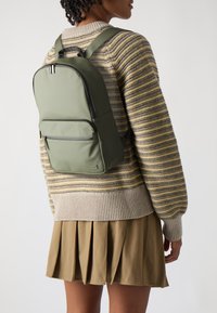 Mochila de nailon verde con una forma estructurada, bolsillo frontal con cremallera y detalles en cremallera negra, llevada sobre un suéter a rayas y una falda plisada.