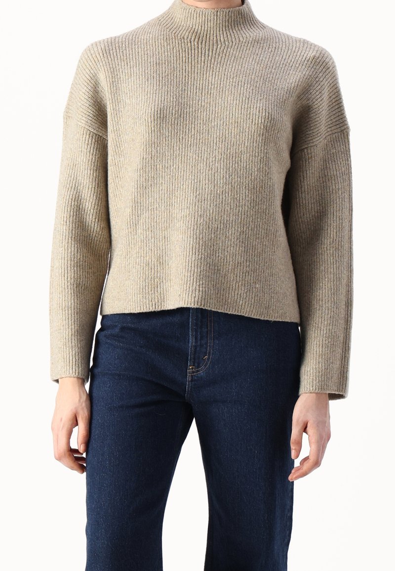 Pull en maille côtelée beige avec col montant et épaules tombantes, présentant une longueur raccourcie et une coupe décontractée, associé à un jean en denim foncé.