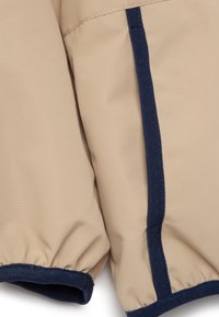 Beige stof met marineblauwe accenten, met elastische manchetten en een visgraatstructuur langs de naden. Close-up van een mouw.