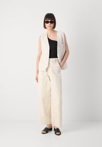 Beige mouwloze vest met knopen, zwarte getailleerde top, brede crèmekleurige broek en zwarte open teen schoenen; model draagt smalle zonnebril.