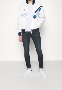 Muž oblečený v bílo-modré vzorované zipové bundě, bílem tričku, tmavých skinny džínách a bílých teniskách stojí na bílém pozadí.