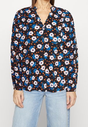 Kvinde iført en sort bluse med hvide og blå blomsterprint og orange prikker, kombineret med lyseblå jeans.