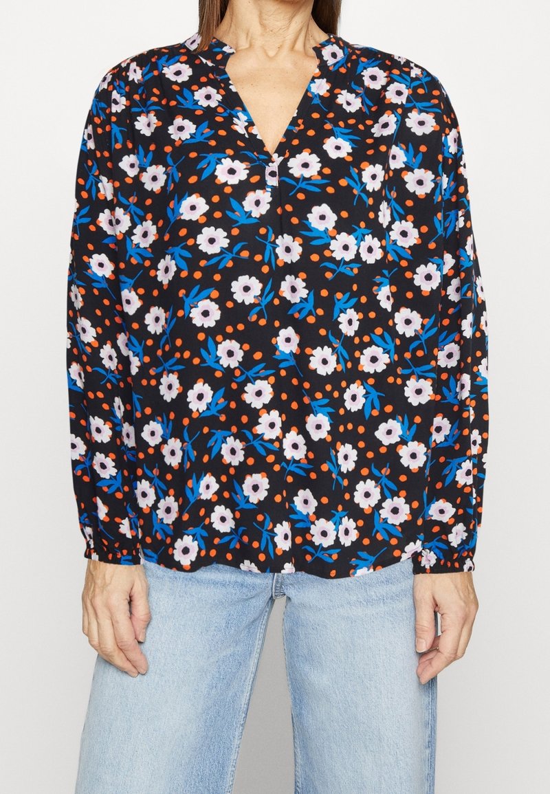 Femme portant un chemisier noir avec des motifs floraux blancs et bleus et des points orange, associé à un jean bleu clair.