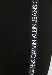 Černé legíny s bočním pruhem a nápisem "CALVIN KLEIN JEANS" v bílé barvě, vyrobené z měkkého materiálu a s přiléhavým střihem.