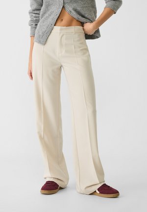 Cremefarvede bukser med vide ben, båret med en grå cropped cardigan og bordeaux sneakers, der viser mellemgulvet og en afslappet positur.