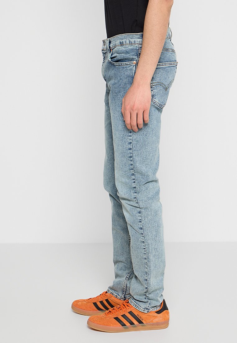 Jeans de denim azul claro con un corte recto, que cuentan con cinco bolsillos, pasadores para el cinturón y un acabado desgastado. Combinados con zapatillas naranjas y negras.
