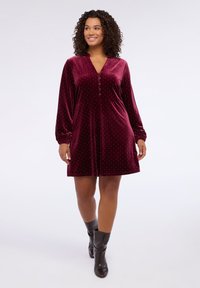 Abito in velluto bordeaux con scollatura a V, maniche lunghe e dettagli con bottoni. Presenta una forma aderente che si allarga verso il basso e accenti di strass sparsi.