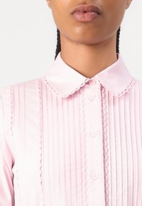 Blusa rosa com gola, pregas verticais, detalhes em renda ao longo da gola e da pala, e pequenos botões na frente.