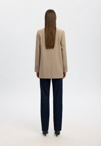 Beige skräddarsydd jacka med avslappnad passform, texturerad tyg, dubbelknappade manschetter, ihop med mörkblå raka jeans och svarta klackar.