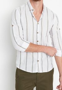 Valkoinen pellavapaita, jossa on pystysuuntaiset harmaat raidat, napillinen kaulus ja käärittävät hihat. Paidassa on puiset napit ja rento istuvuus.