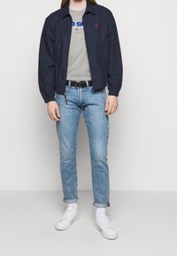 Námořnická lehká bunda s elastickými manžetami; šedé tričko s grafikou a červeným a modrým textem; světle modré džíny; bílé tenisky; ležérní vzhled.