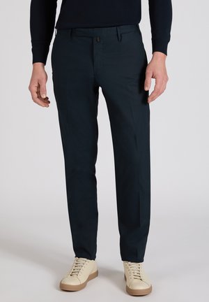 Mężczyzna ubrany w dopasowane ciemnogranatowe spodnie i kremowe trampki, stojący na tle jednolitego jasnego tła.