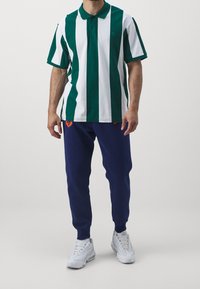 Grün-weiß gestreiftes Poloshirt mit Kragen, kombiniert mit navyblauem Sweatpants mit orangefarbenen Akzenten und weißen Sneakers.