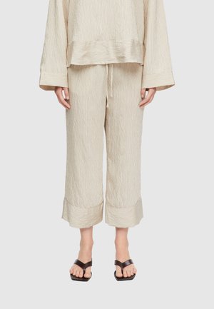 Beżowe, krótkie spodnie o szerokich nogawkach z subtelnymi pionowymi paskami i podwiniętymi mankietami, noszone z dopasowaną bluzką z długim rękawem oraz czarnymi sandałami z kwadratowym noskiem.