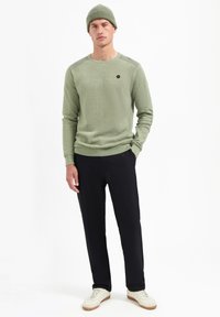 Lichtgroene gebreide trui met ribbelaccenten, gecombineerd met een zwarte broek en witte sneakers. Wordt aangevuld met een bijpassende groene muts.